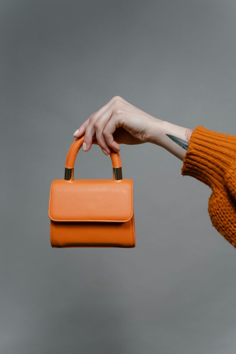 A stylish orange leather handbag is held by a tattooed hand with a knitted orange sweater sleeve.