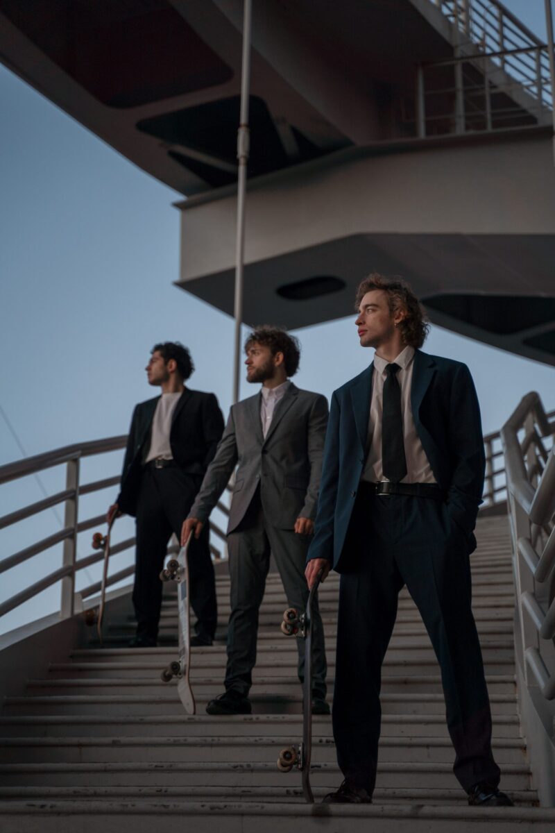 Three men in suits holding skateboards on modern architectural stairs during daytime.