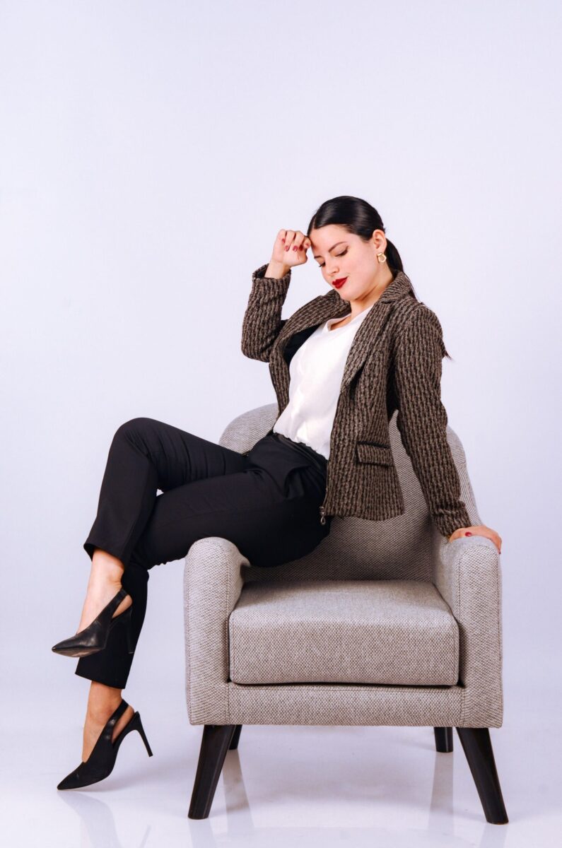 Elegant woman posing in a studio sitting on a gray armchair, dressed in a professional outfit.