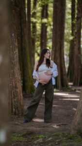 Pregnant woman standing among tall trees, embracing her belly in a peaceful forest setting.