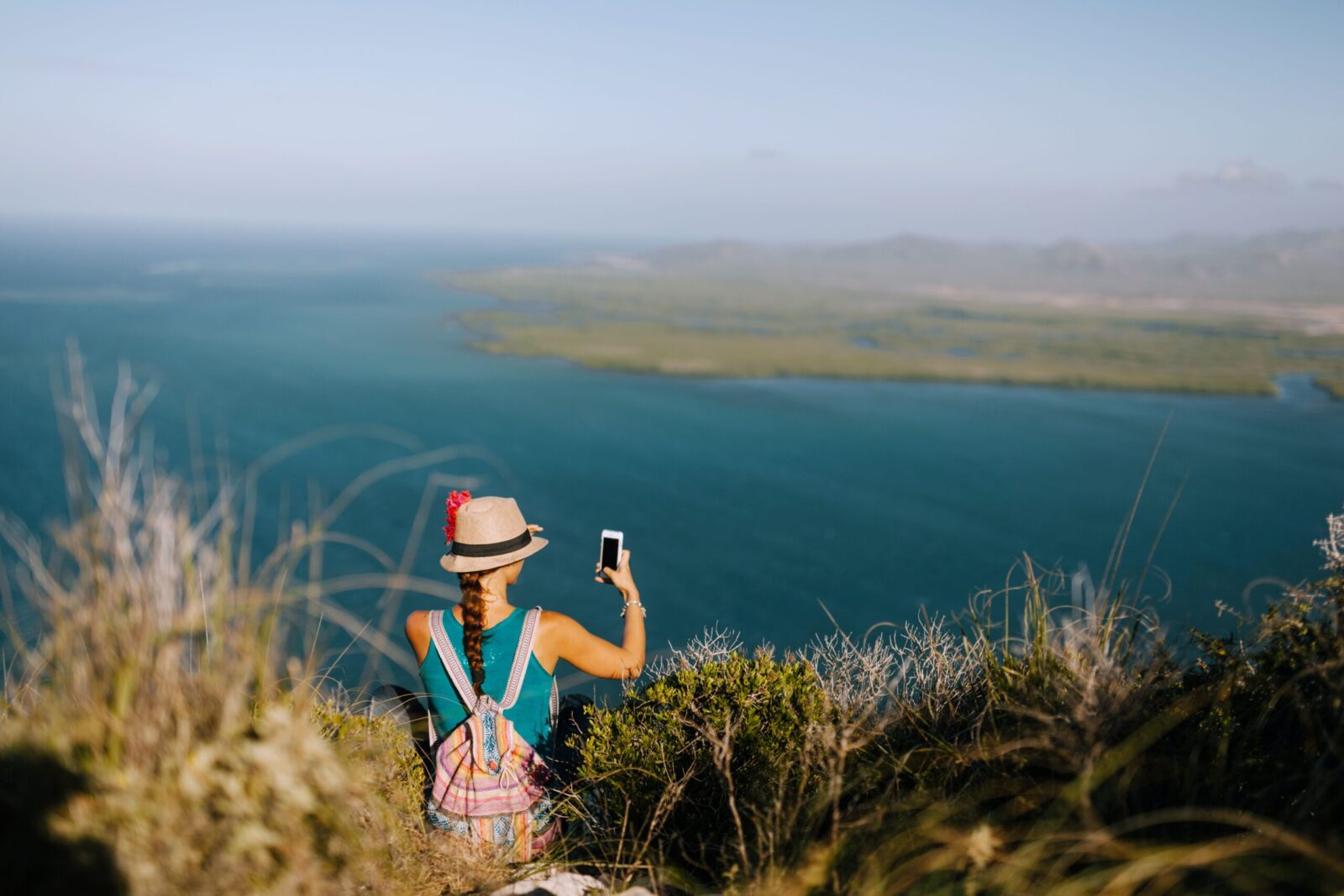 Back view of unrecognizable female traveler taking photo of foggy ocean on cellphone while resting on grass mount